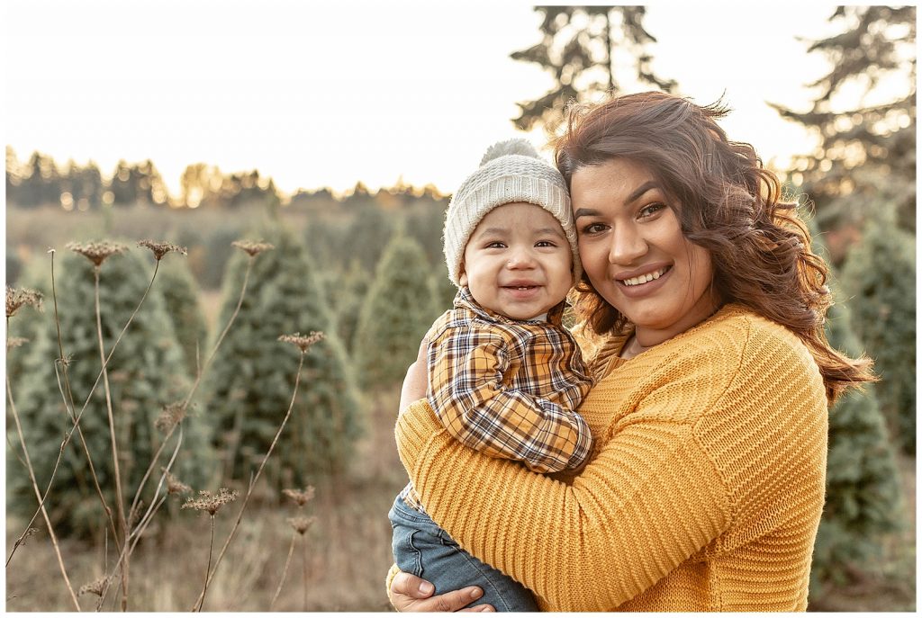 Mom in gold sweater and one year old son in gold plaid shirt and cream hat stand closely together in field of Christmas trees for family photos.