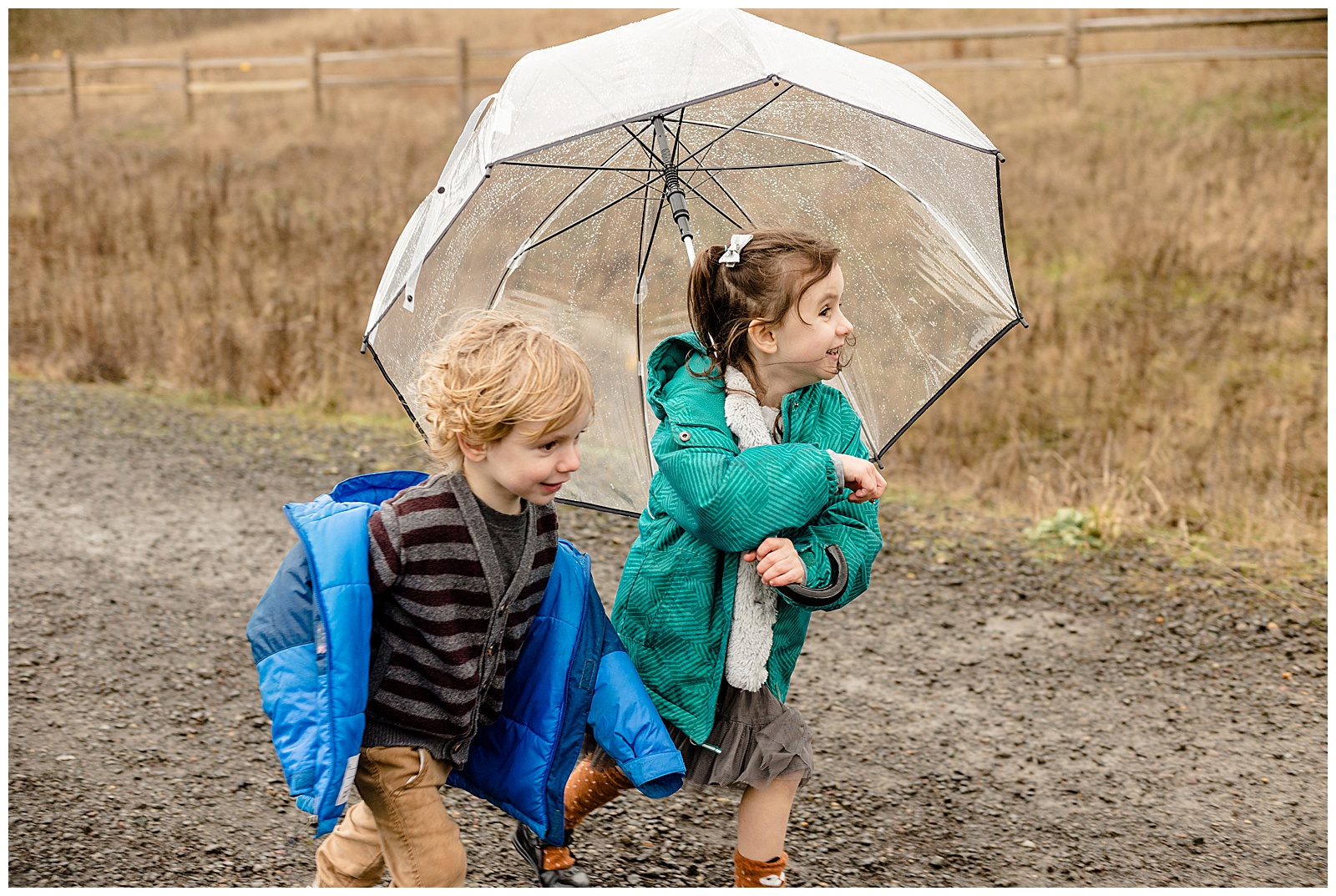 3 year old boy and 5 year old girl running with rain coats and clear umbrella