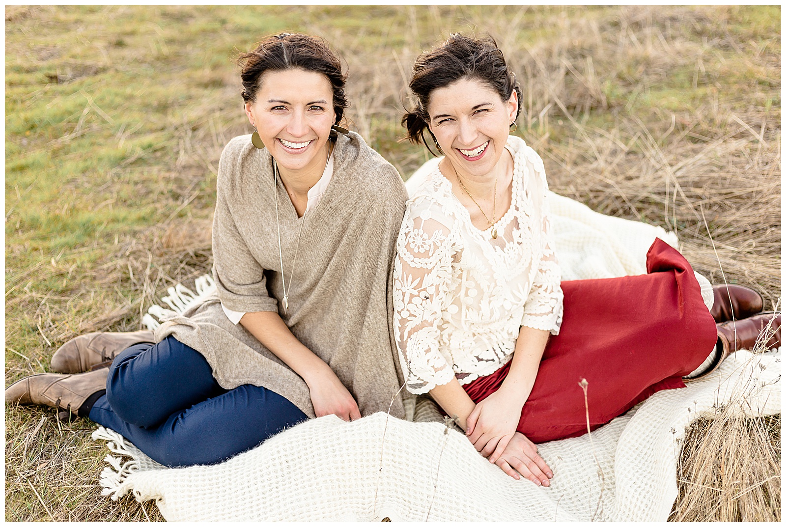 2 Sisters in their 20's sitting on a biege blanket out in nature both smiling at the camera. 