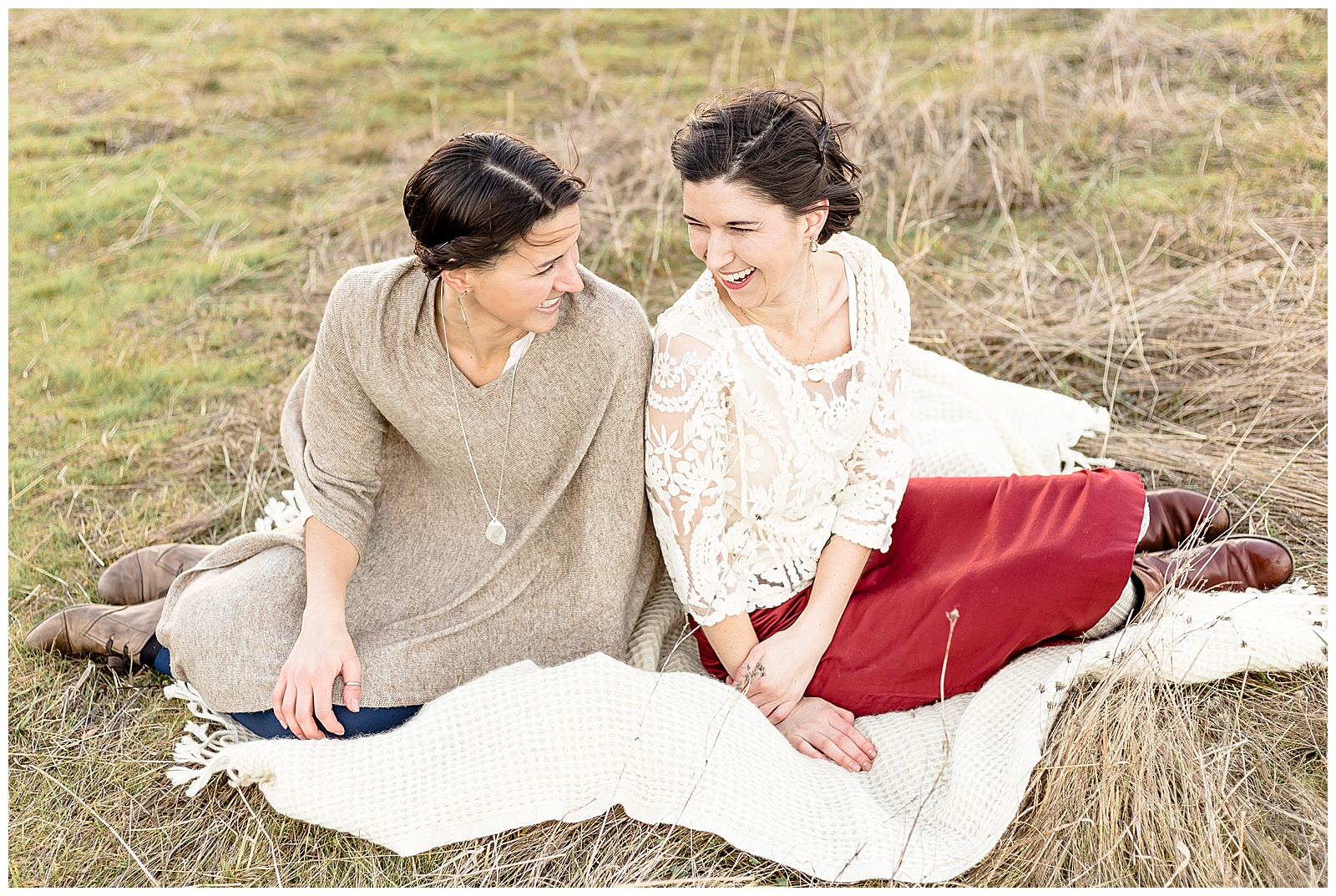 2 Sisters in their 20's sitting on a biege blanket out in nature both smiling at the camera. 
