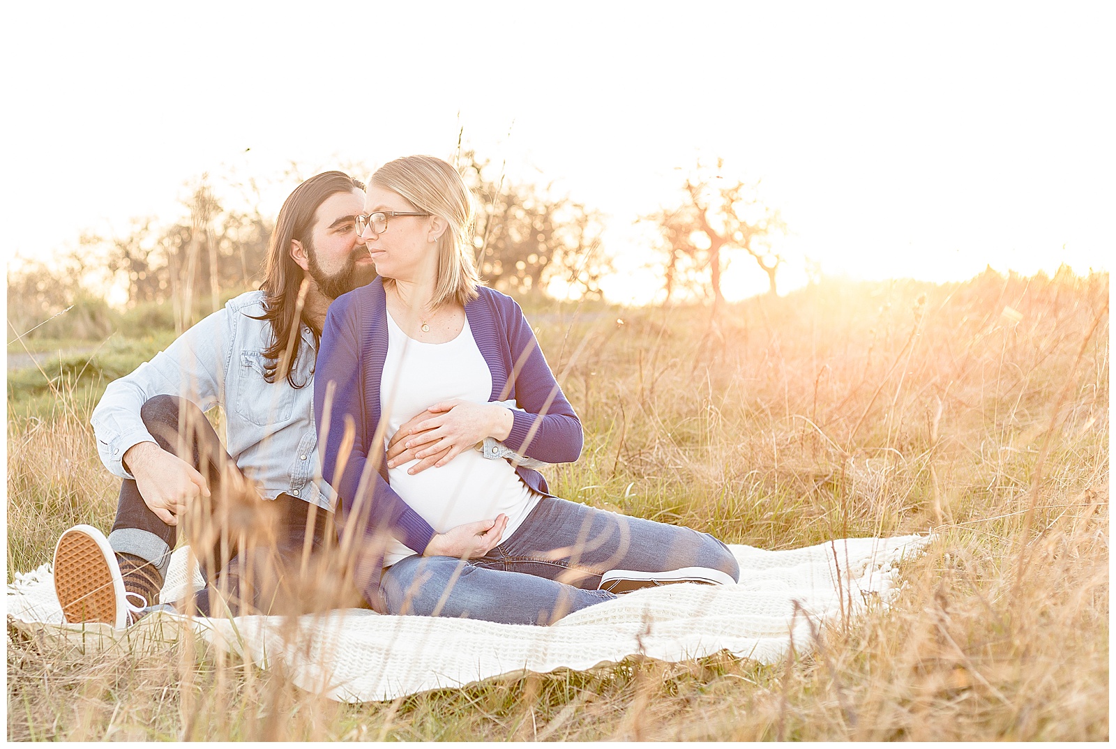 Pregnant Mom with white shirt and blue sweater and dad in denim sitting on a blanket in nature at sunset