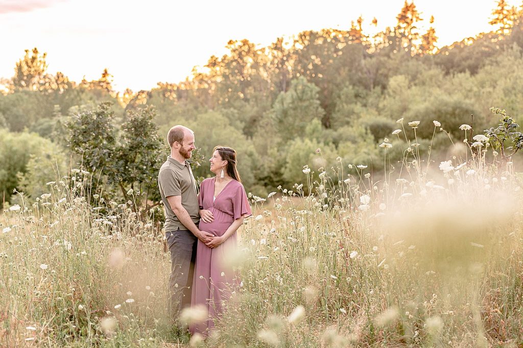 man in olive green shirt and pregnant woman in a mauve dress standing next to each other and looking at one another in the middle of wildflowers