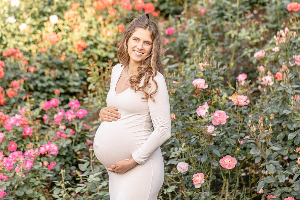 White skinned woman in white dress holding pregnant belly in front of pink roses