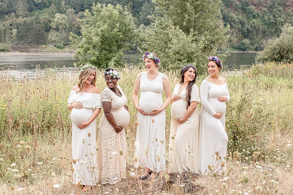 diverse group of five pregnant woman out in nature in beautiful white and cream dresses looking at each other and laughing