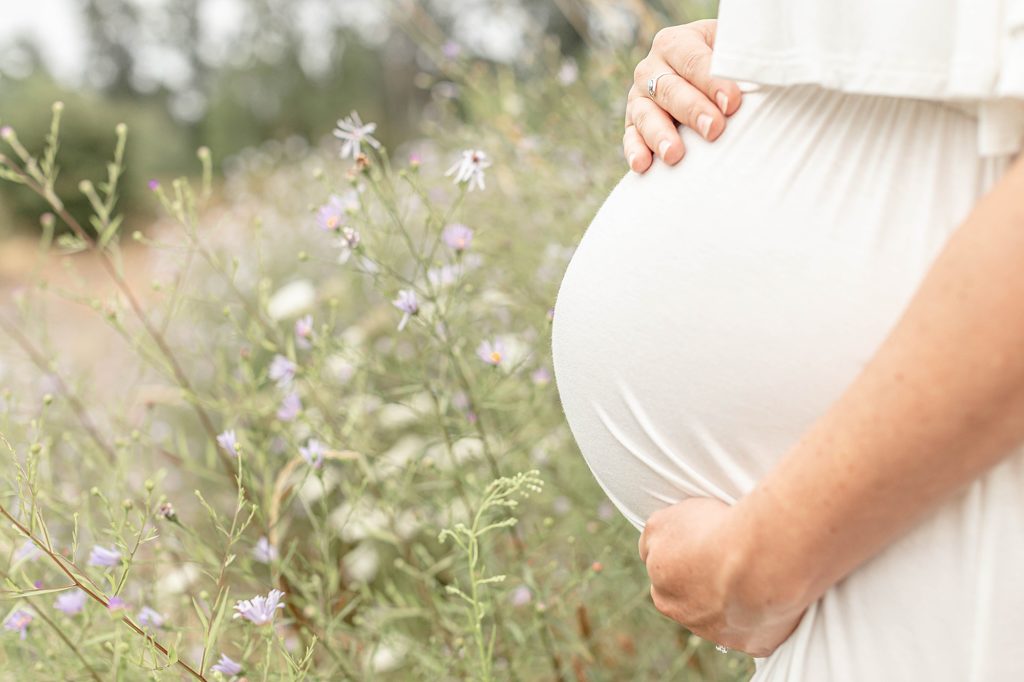 Light skinned woman holding pregnant belly surrounded by wildflowers
