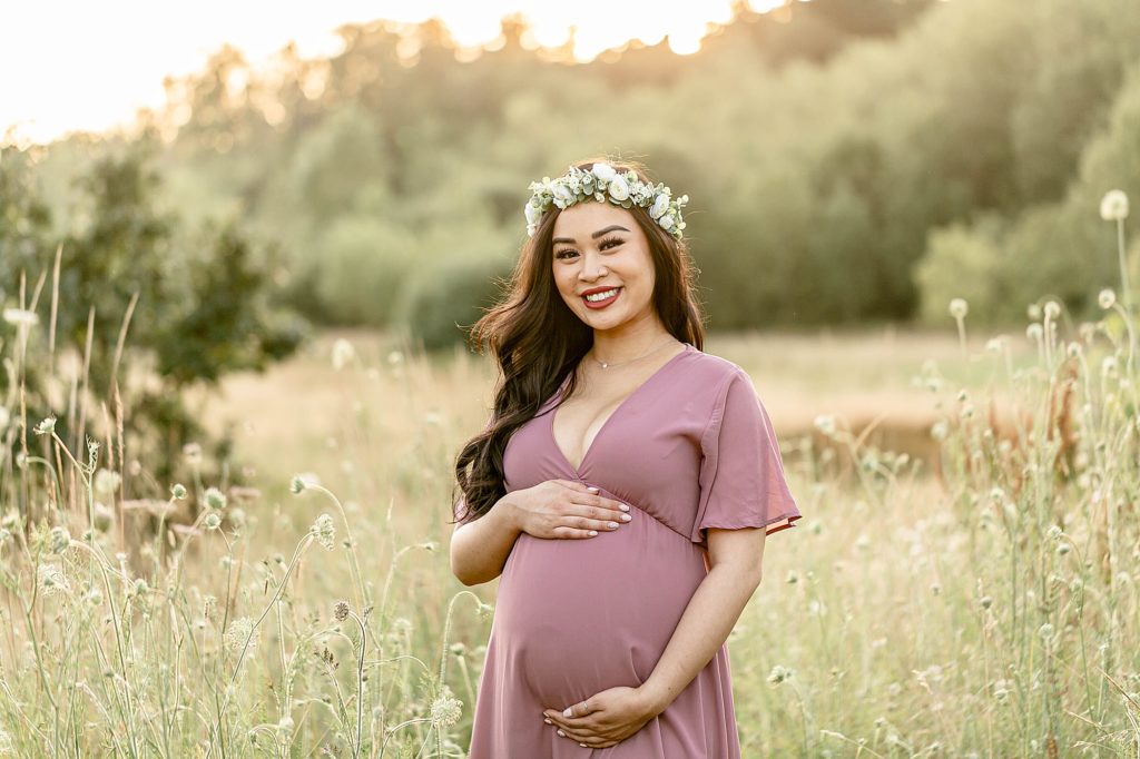 Pregnant woman wearing flower crown and long flowy dress out in nature with beautiful sunset