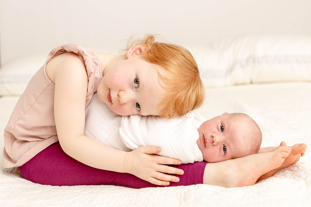 light skinned toddler girl with red hair sitting on bed with newborn baby brother in her lap both looking at the camera