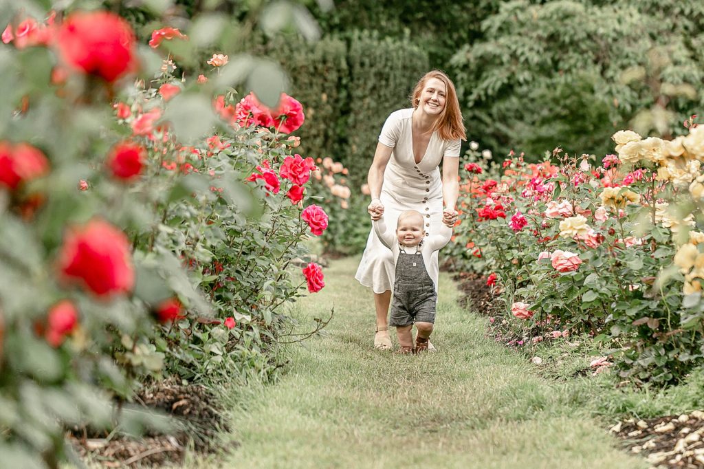 Light-skinned woman holding baby boy hands walking in the rose gardens