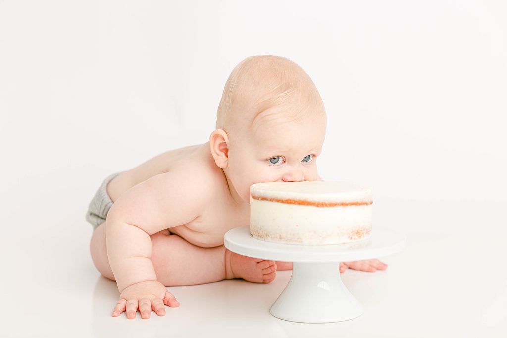 Light-skinned baby shoving head into a naked cake in a white photography studio