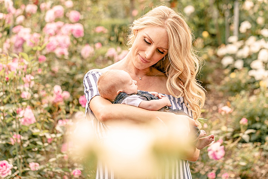 Light-skinned woman holding newborn baby framed by rose in the rose gardens