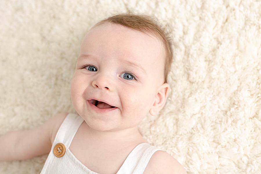 light-skinned six month old baby boy in white romper on a cream rug smiling at the camera - portland baby photographer