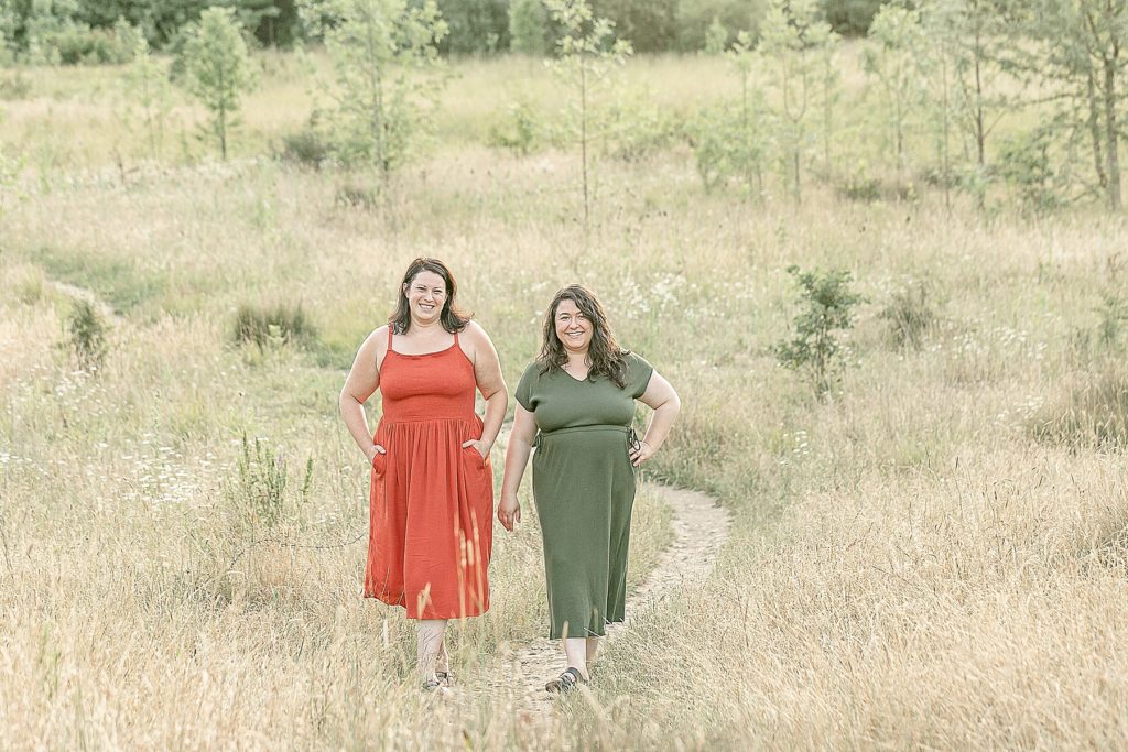 Two light skinned women walking out in nature in long grasses. One is wearing an orange tank top dress and the other is wearing an olive green short sleeve dress