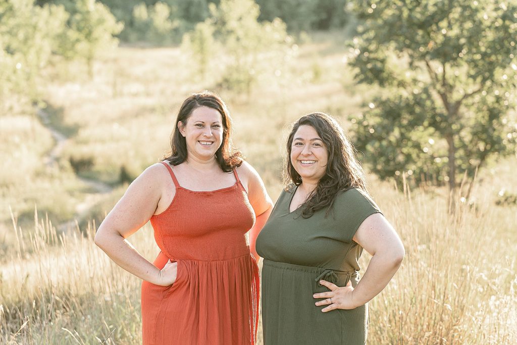 Two light skinned women out in nature. One is dressed in an orange summer dress and the other is dressed in an olive green dress. They are both doulas in SW Portland Oregon.