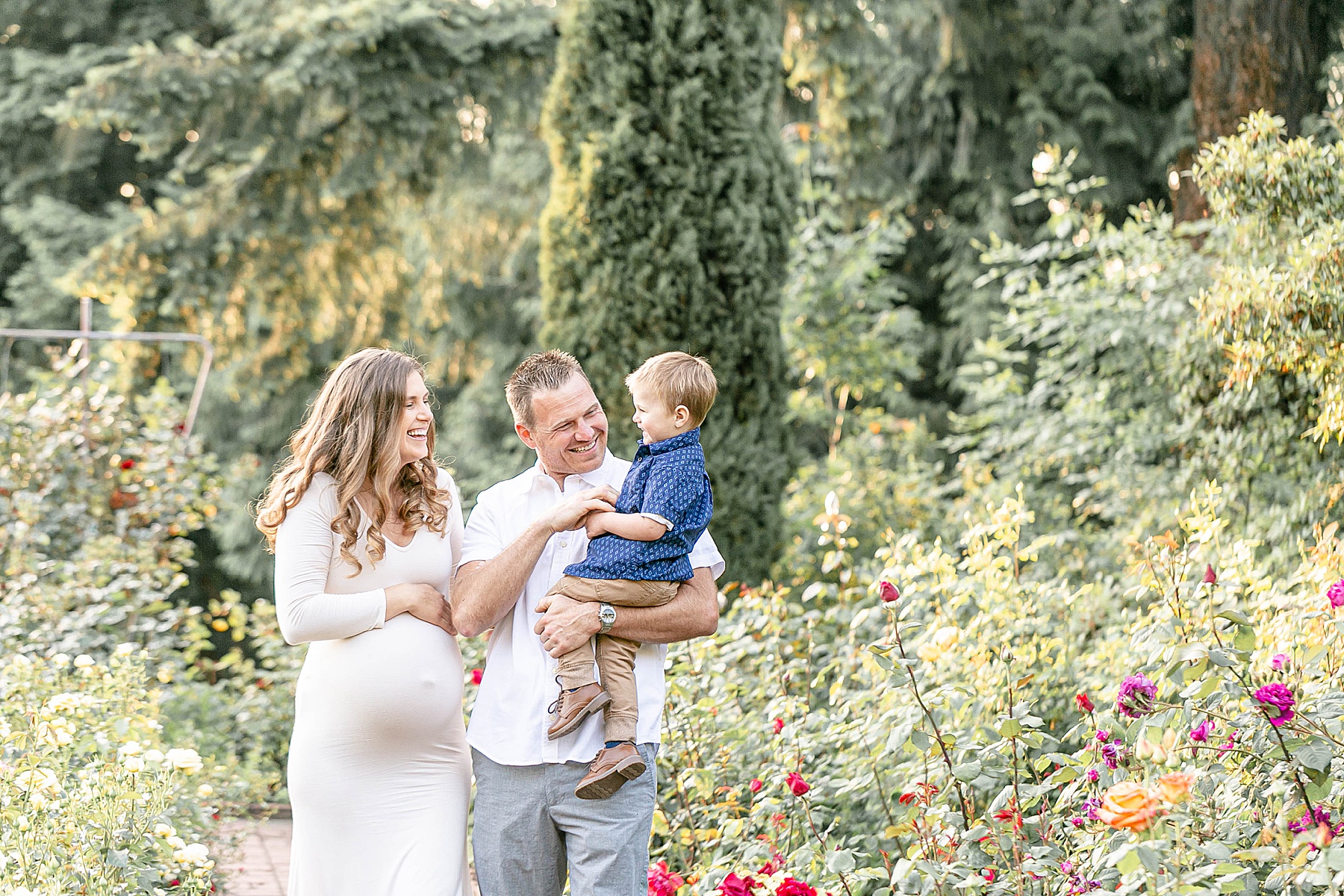 pregnant woman in white dress with man in white shirt & grey pants holding little boy in blue shirt and khaki pants giggling together in the Portland International Rose Test Gardens