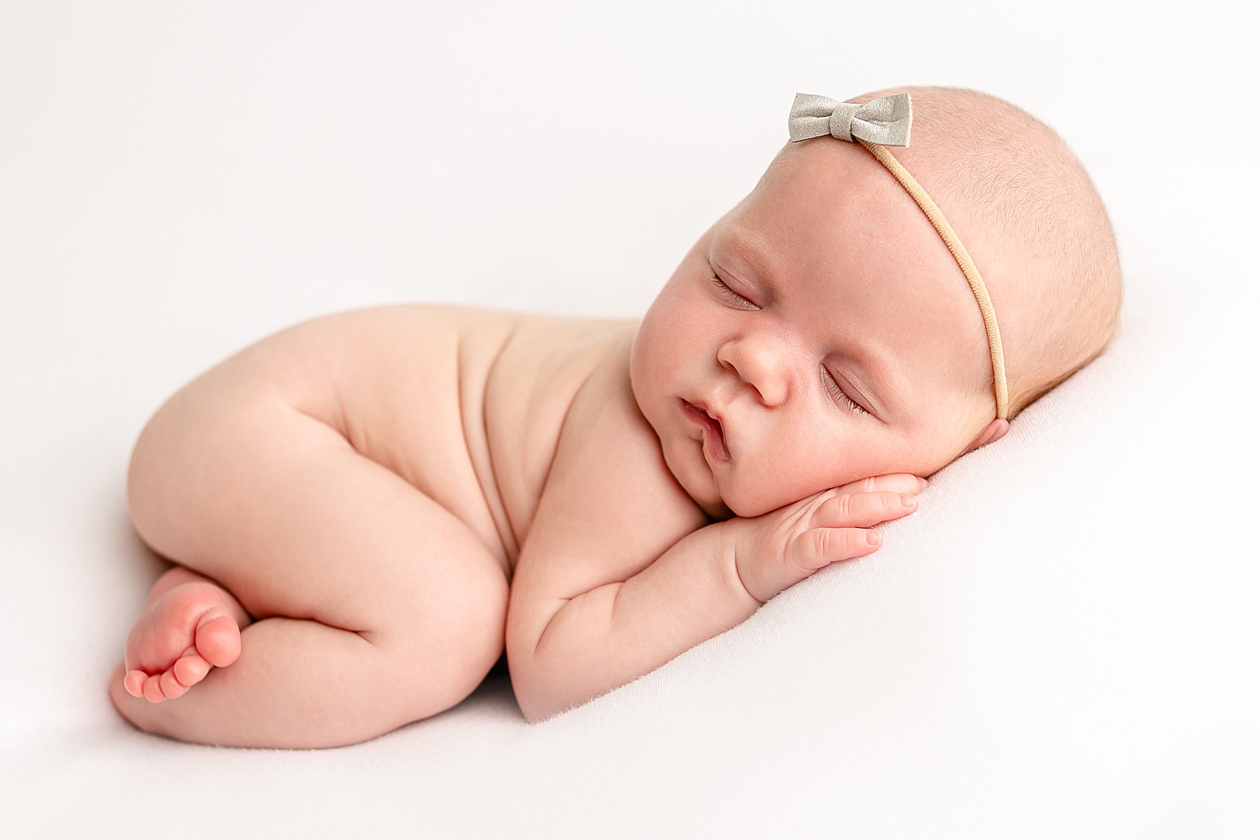 light-skinned baby curled up on white backdrop with minimalist grey bow on head - light and airy newborn photos portland oregon