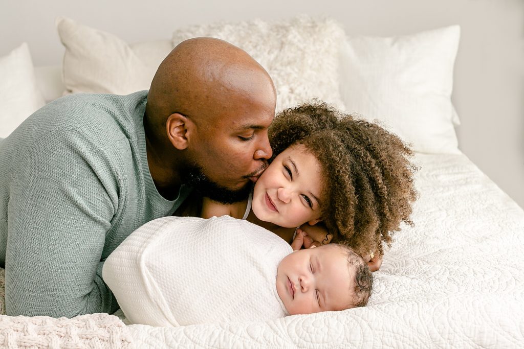 Dad kissing big sister while baby sleeps next to them during newborn portrait session sw portland