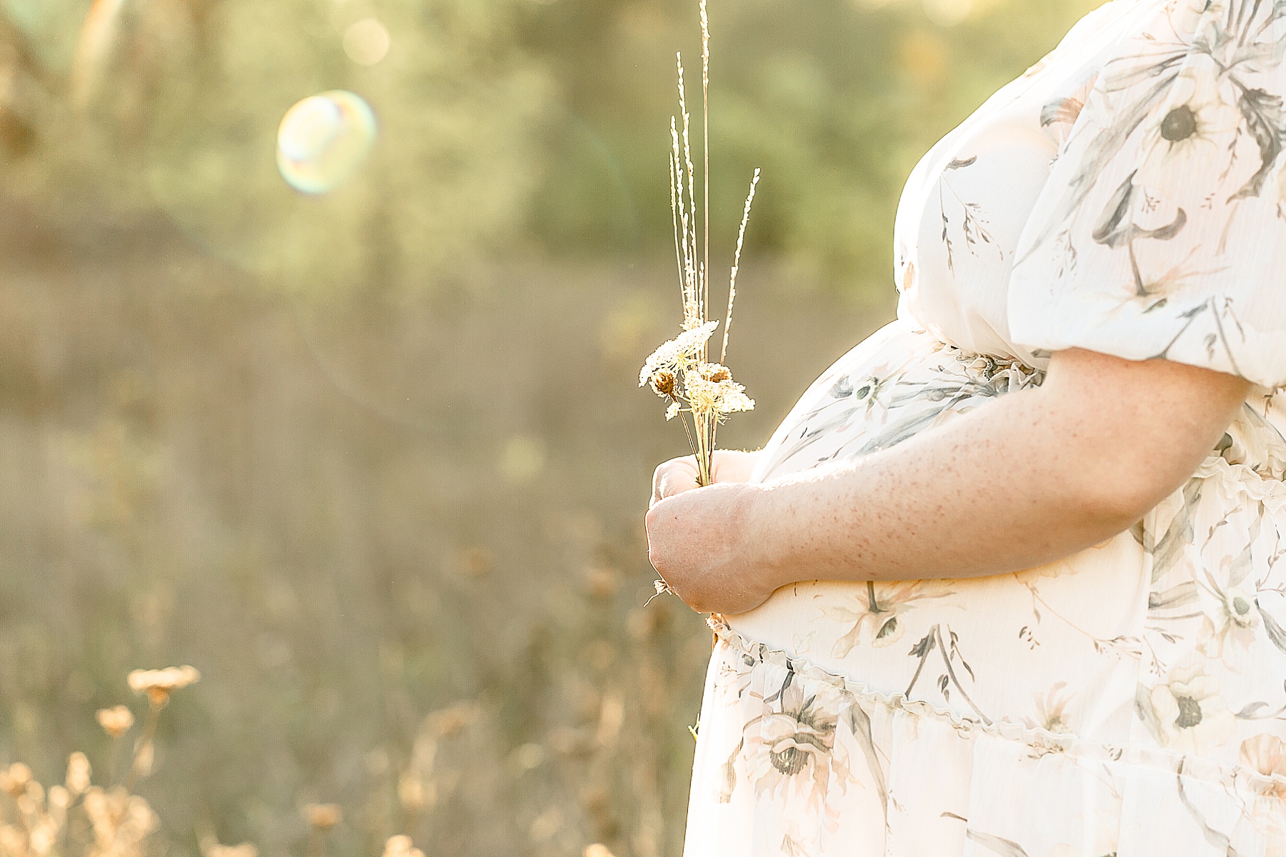 Pregnant woman in white floral dress holding flowers in front of pregnant belly outside