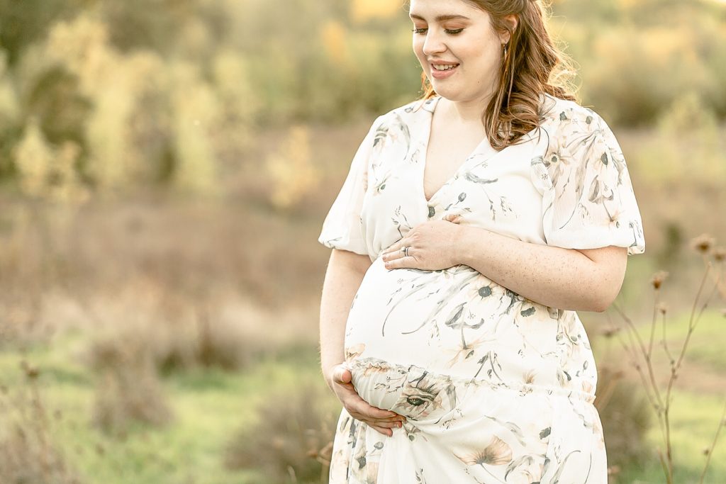 Light-skinned woman in white floral dress holding pregnant belly at baby bump photo session outdoors in nature