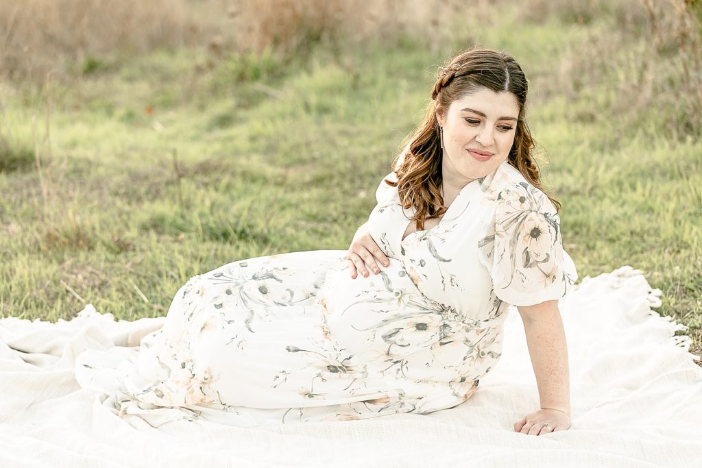 light-skinned woman holding pregnant belly sitting on blanket outside during photo session with best maternity photographer in portland oregon