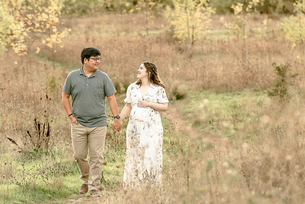 Asian man and white woman dressed in light neutral colors and walking down a nature path looking at each other during their maternity photography session in SE Portland Oregon
