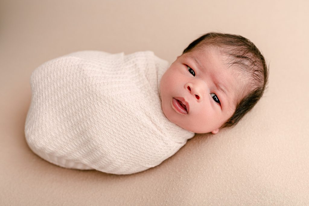 Newborn baby wrapped in cream swaddle on beige backdrop wide awake