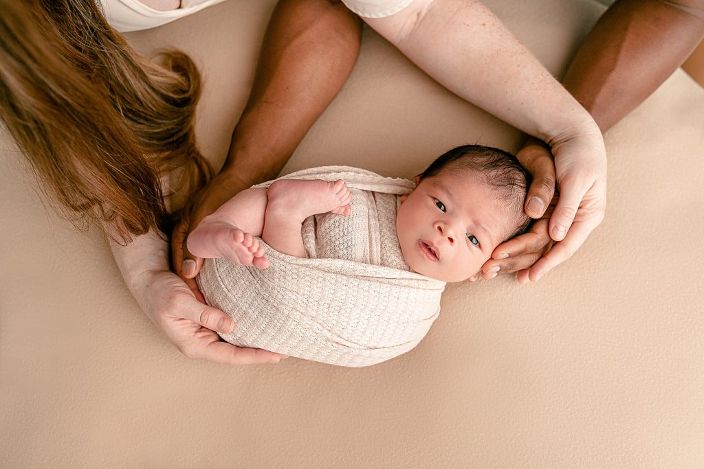 Mom and Dads arms holding baby with beige backdrop newborn photos