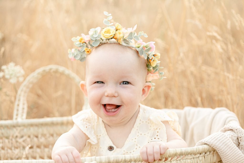 light-skinned seven month old baby with a flower crown sitting in a moses basket in a field of tall golden grasses during family milestone photo session