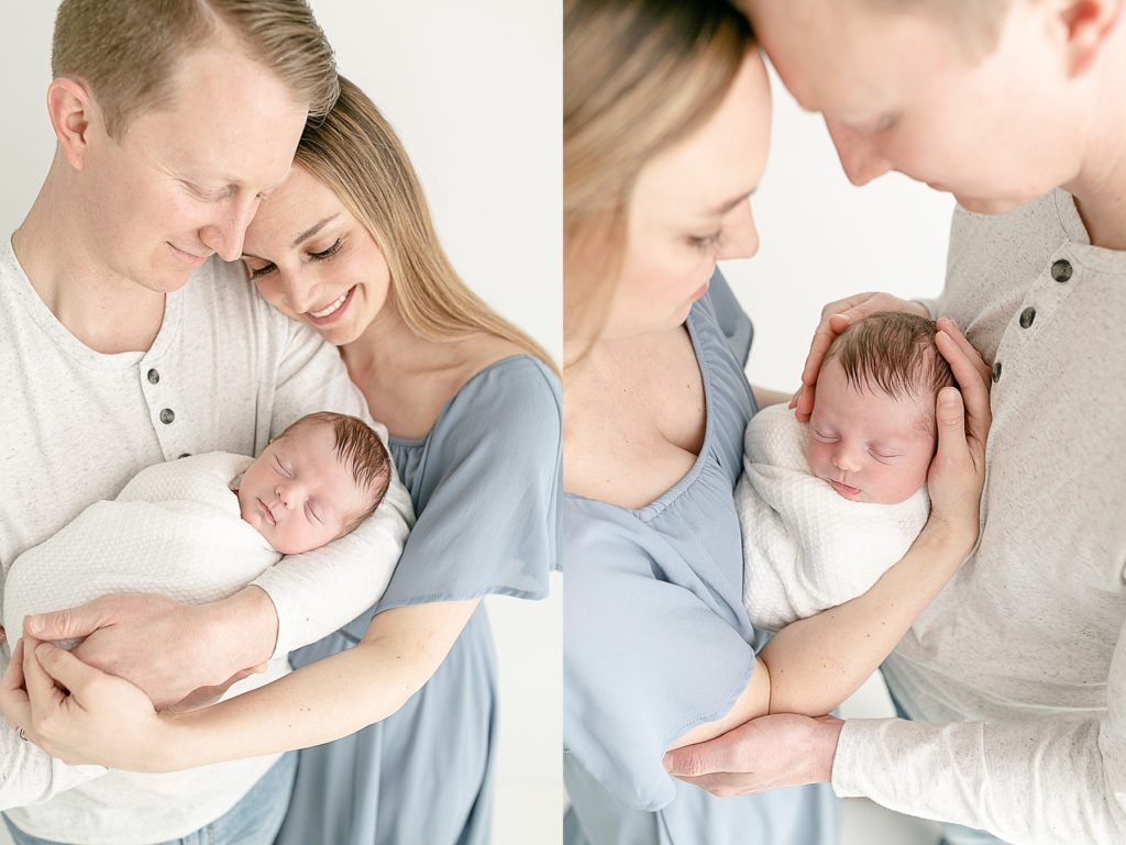 Mom dressed in blue dress, dad in grey shirt holding newborn baby wrapped in white blanket