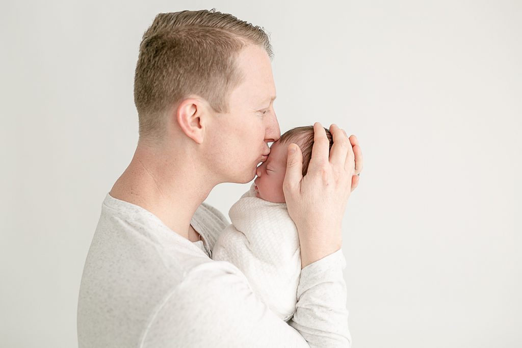 dad dressed in white and baby swaddled in white dad kissing baby's forehead