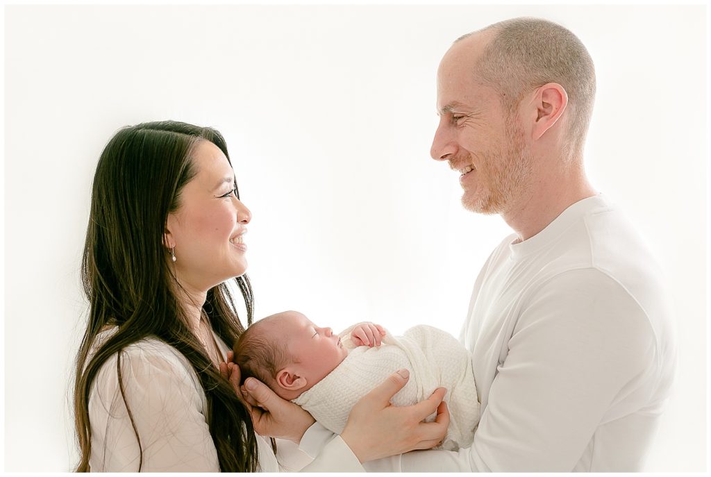 mom and dad wearing light, neutral colored outfits holding baby at newborn portraits