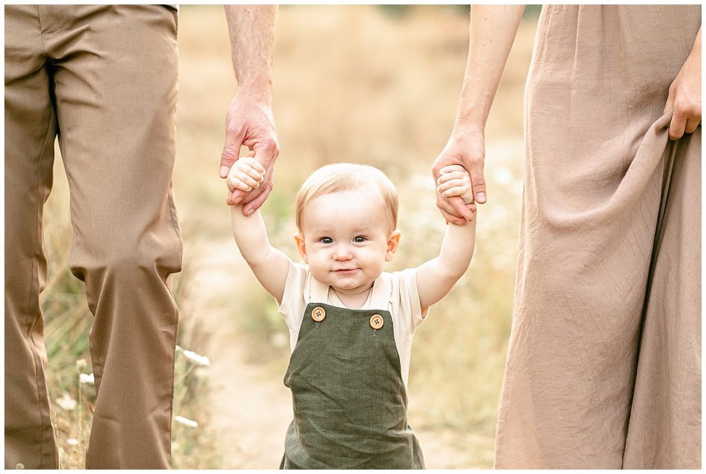 one year old boy in green linen jumper holding parents hands and looking at the camera - portland family photos