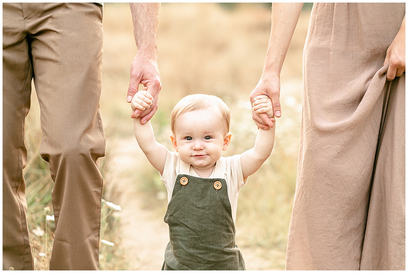 one year old boy in green linen jumper holding parents hands and looking at the camera