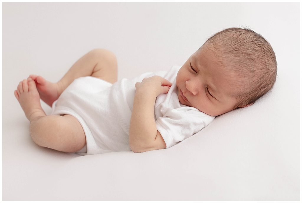 light-skinned newborn baby in white onesie on white backdrop sleepy at newborn photography session.