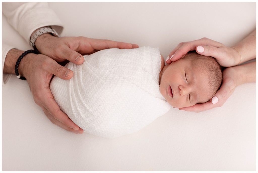 Newborn baby swaddled in white on white backdrop with parents hands on him.