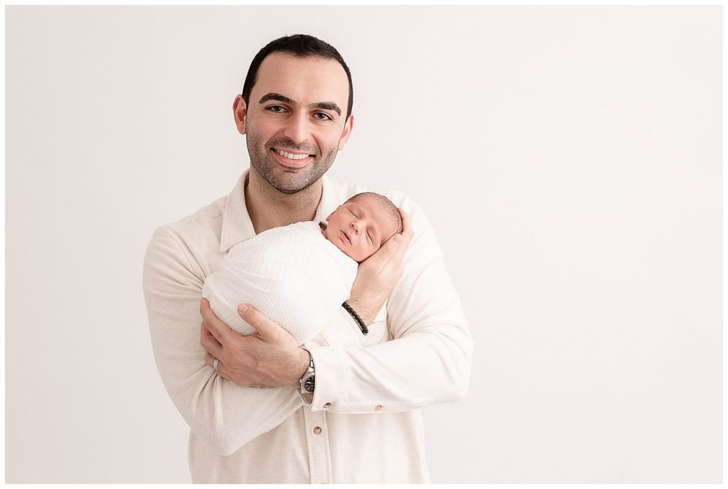 Dad dressed in cream button-down shirt holding baby and smiling at the camera.
