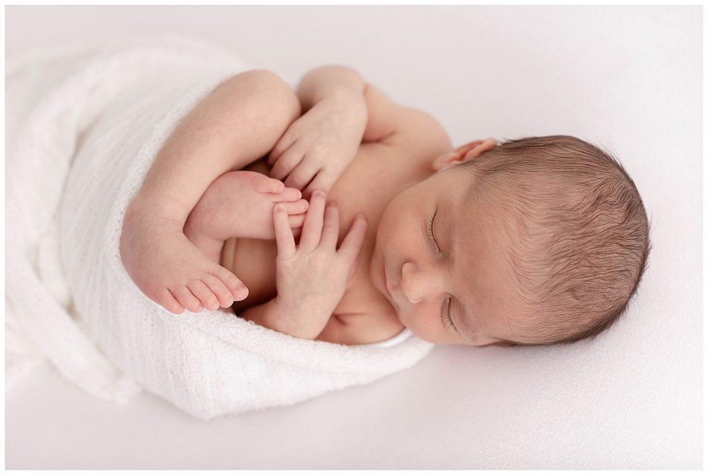 light-skinned newborn baby sleeping with white fabric wrapped around his body. Hands and Feet visible.