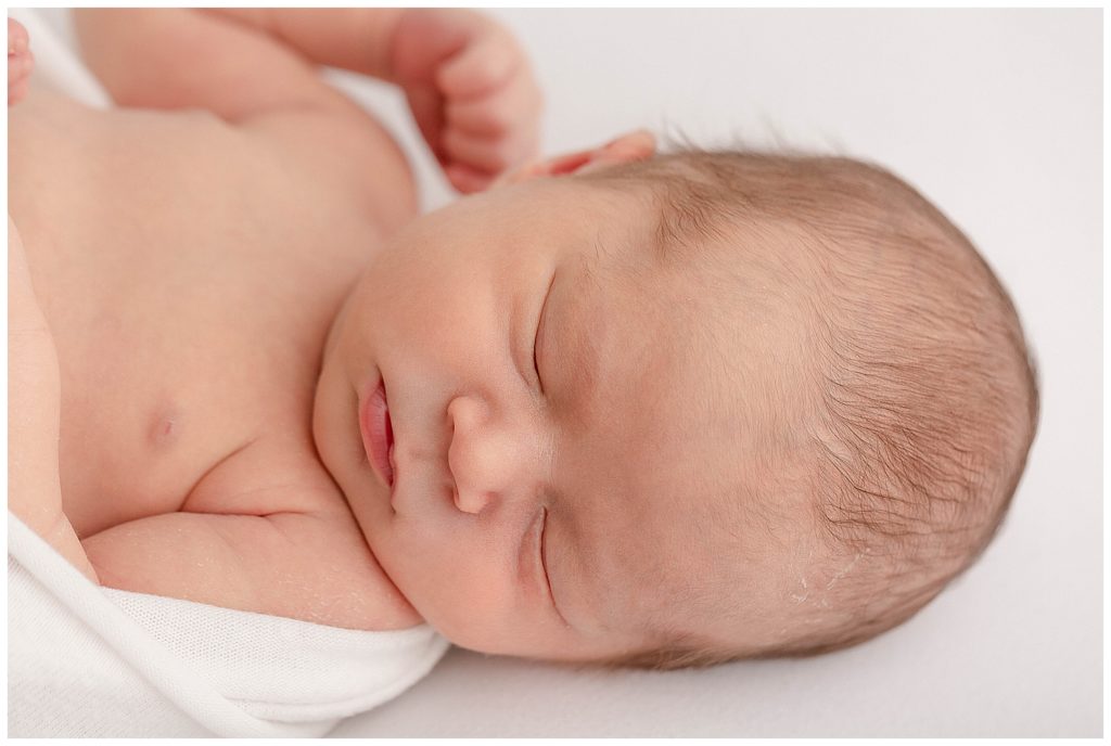 Professional newborn photo of close-up of baby head and chest. baby is laying on a white backdrop with head turned off the shoulder and looking at the camera. Baby is sleeping. 