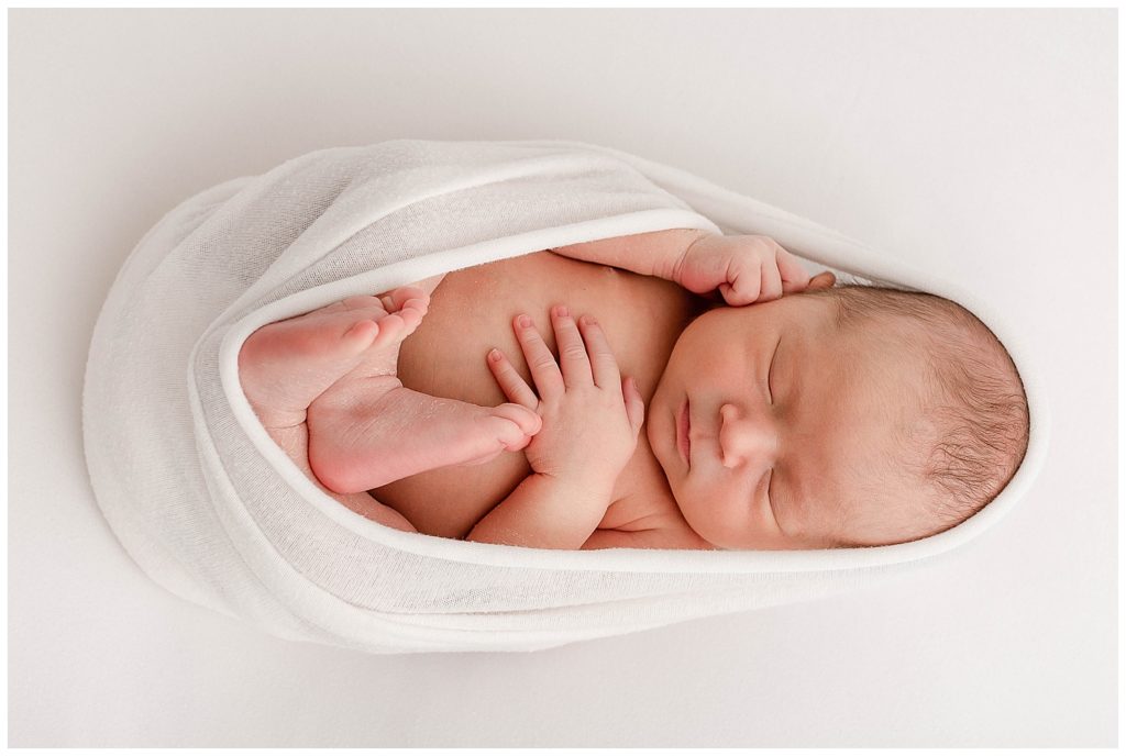 Light-skinned newborn baby wrapped in a white swaddle with some of it slightly over his head. His hands and feet are out of the swaddle. 