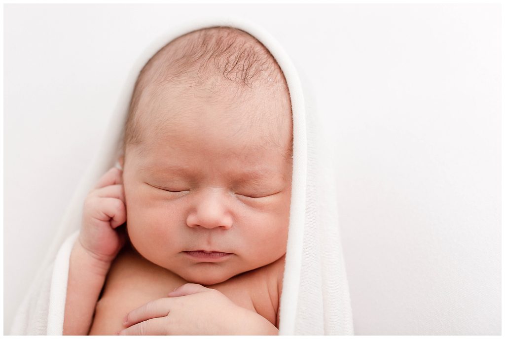 close-up of baby's head and shoulders. light-skinned newborn baby wrapped in white swaddle on a white backdrop with one hand on chest and one hand up by cheek. Posed during his professional newborn photos. 