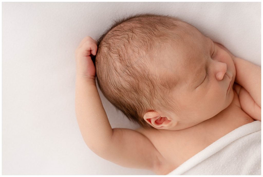 Light-skinned newborn baby laying on white backdrop with arm up by head and has a white blanket over chest. 