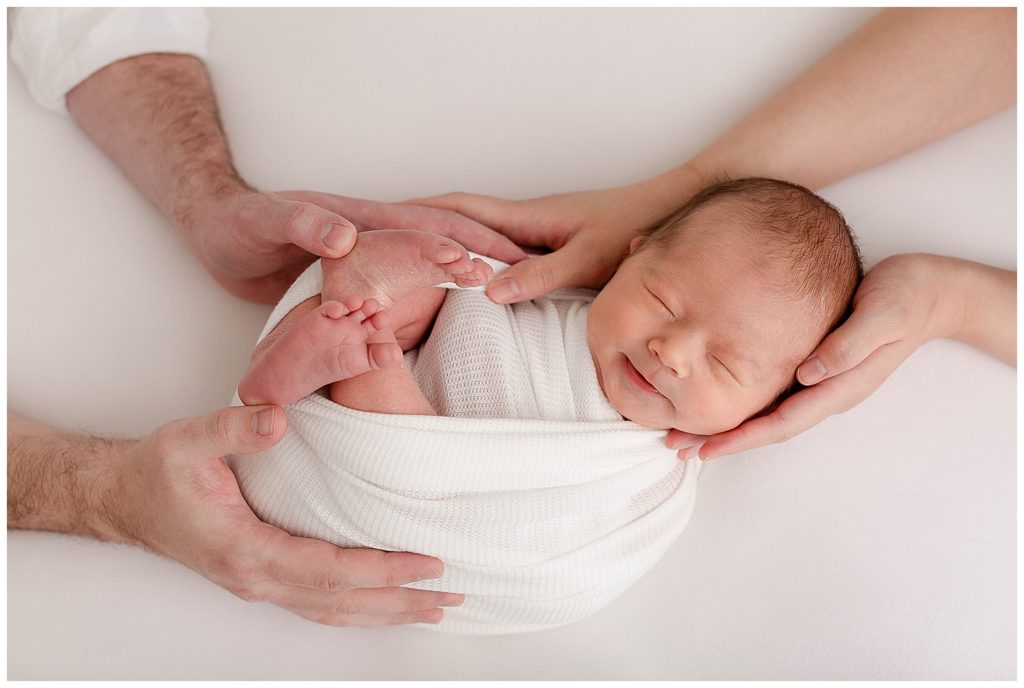 light-skinned baby wrapped in a white swaddle with parents hands on head and at feet. Photo taken during professional newborn photo session