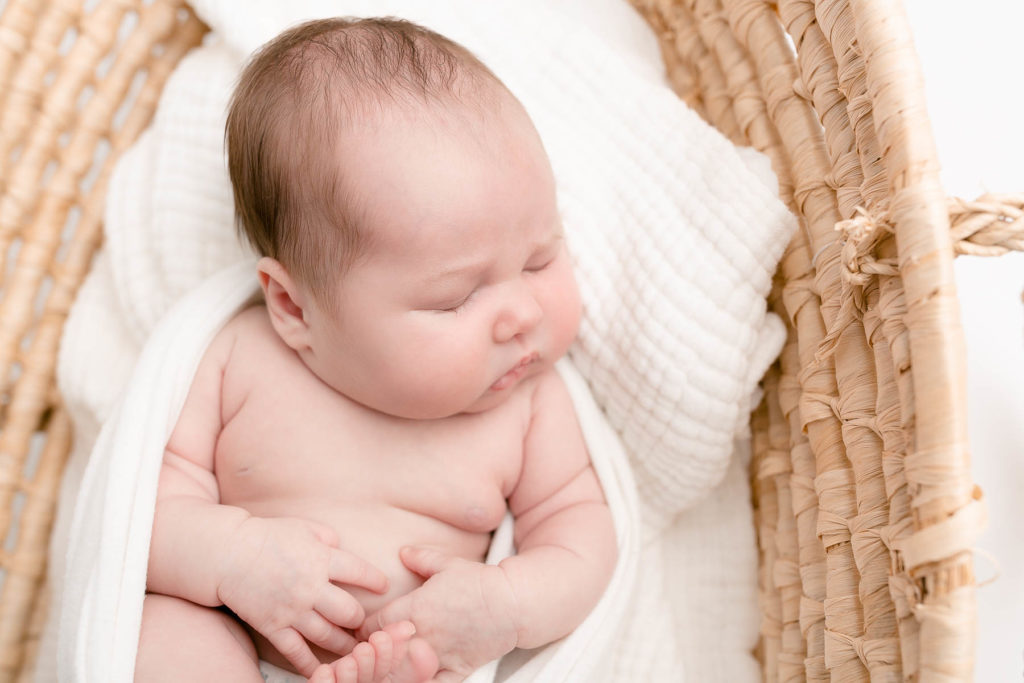 Light-skinned newborn baby sleeping in Moses basket on white blankets at newborn photography session