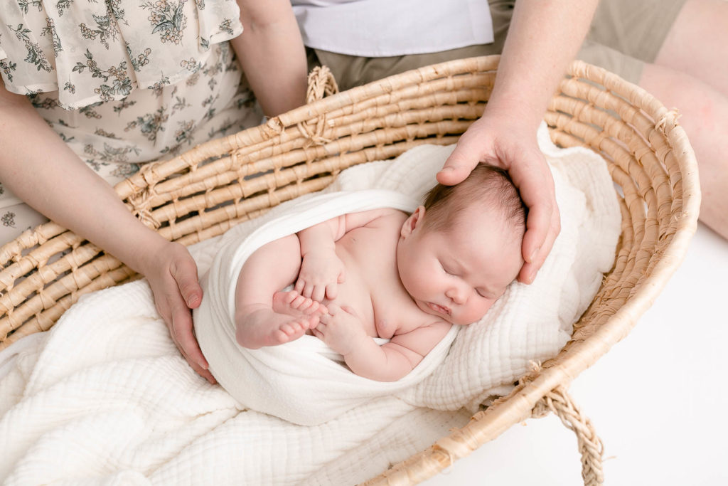 Light-skinned newborn baby wrapped in white laying in Moses basket with parents sitting beside basket. Each parent has a hand on baby. 