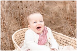 One year old baby girl wearing a pink dress with a white sweater and sitting in a Moses basket at outdoor family photography session. She is laughing off camera and sitting in a field of tall golden grass. 