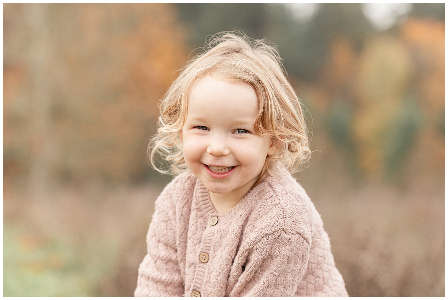 little girl in pink sweater and blonde curls is smiling at the camera.