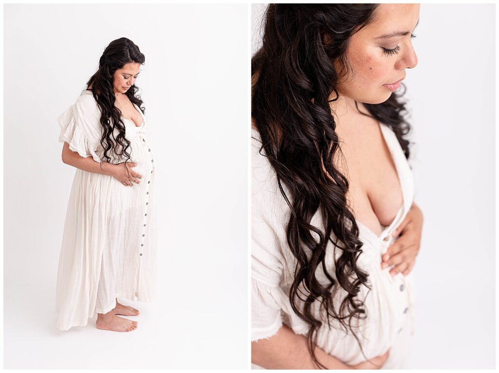 Pregnant woman with long dark curly hair in white flowy gown looking down at her baby bump.