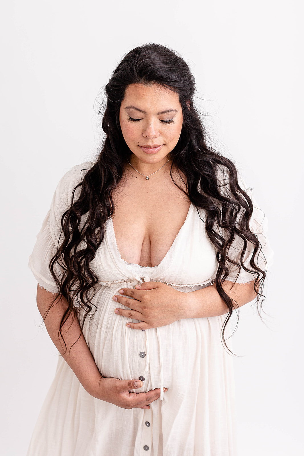 A mother to be with long black hair stands in a studio looking down at her bump
