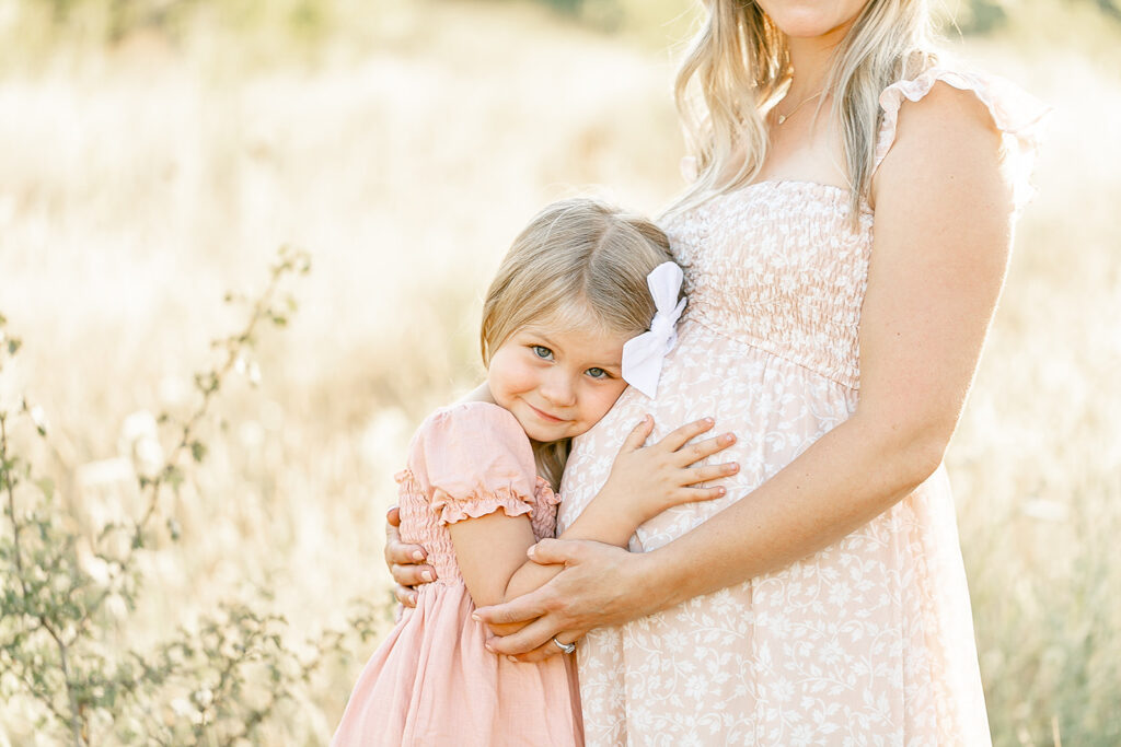 A young girl in a pink dress hugs and rests her head on her mother's pregnant belly