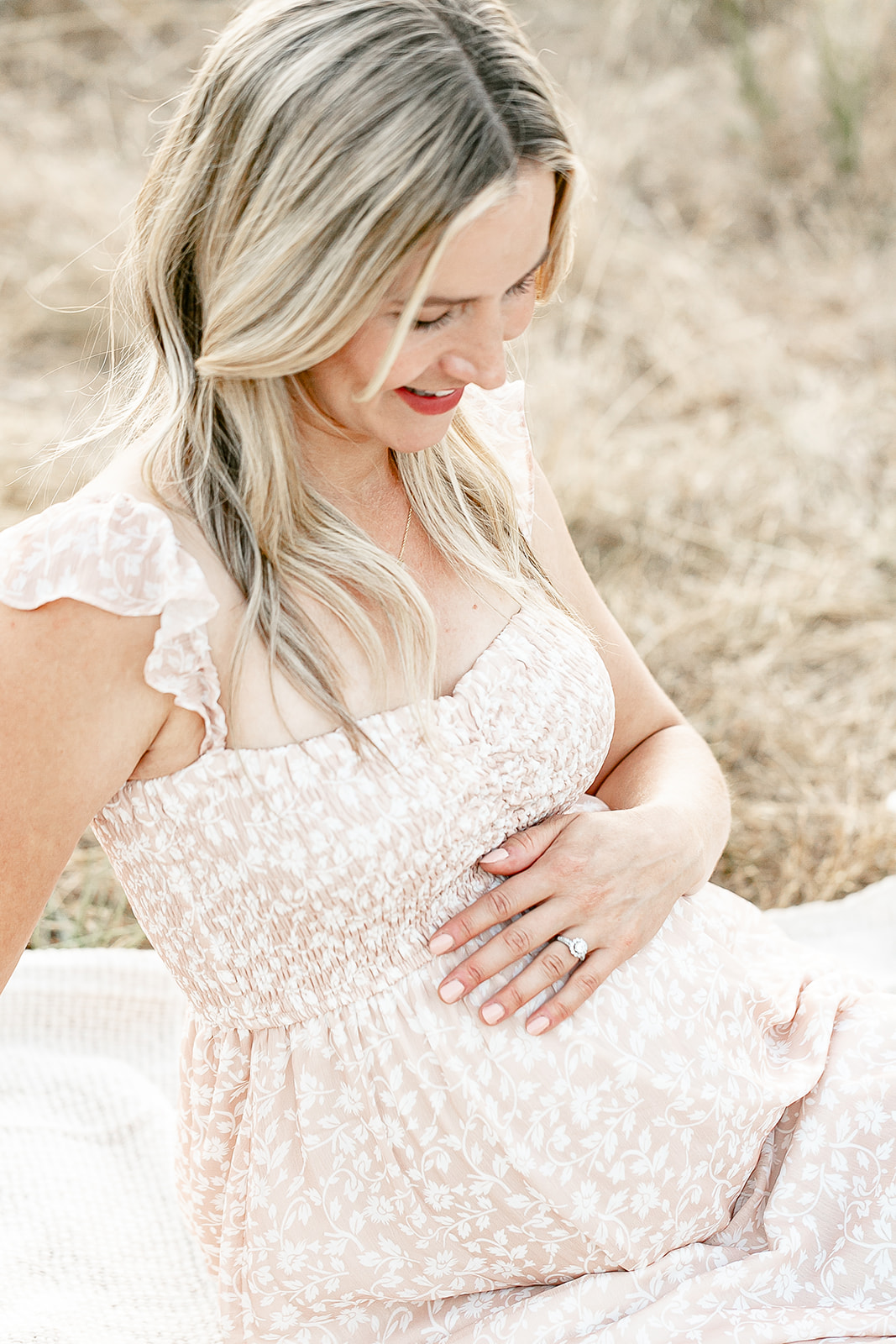 A mother-to-be sits on a blanket in a grassy field while looking down at her bump in a beige dress Portland prenatal yoga