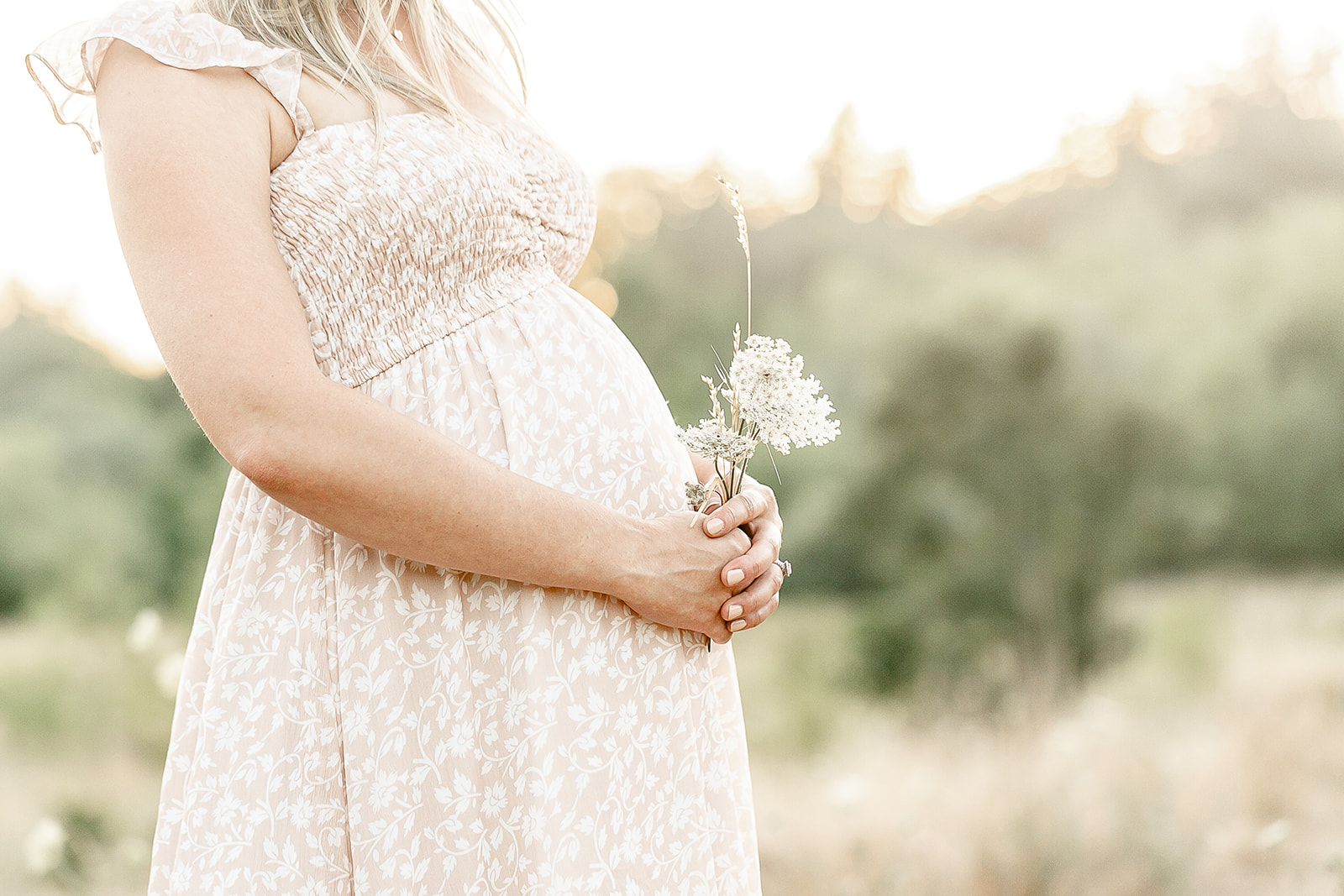 A mother to be stands in a field in a beige floral pattern dress holding small white flowers on her bump Portland prenatal yoga